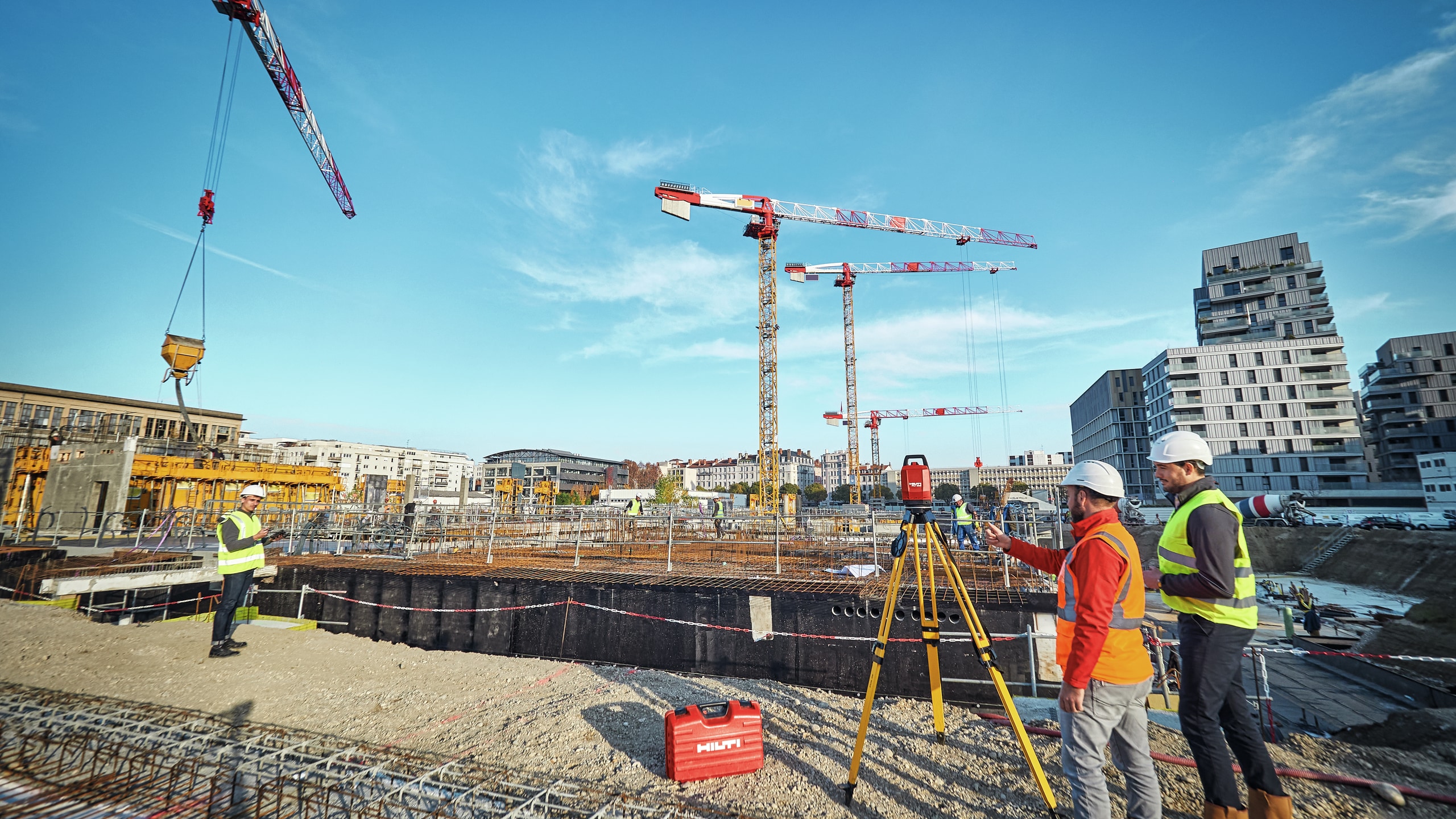 To personer ser på en tablet på en tripod på en byggeplads med kraner i baggrunden