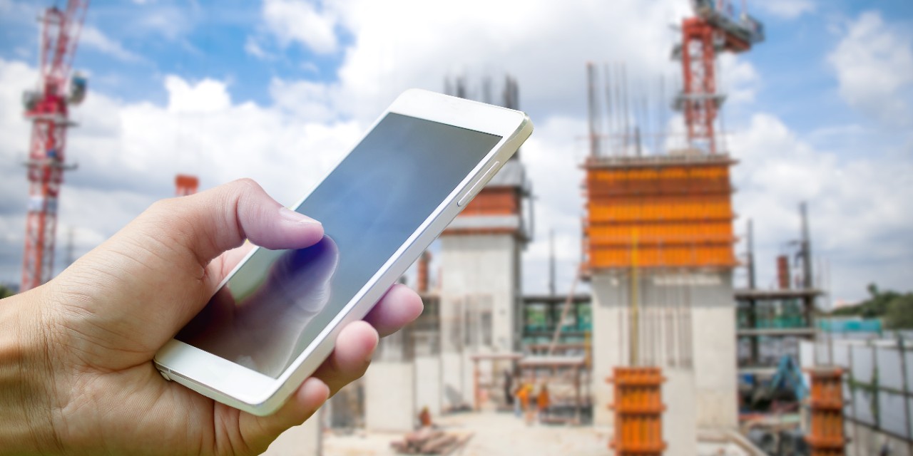 Hand holding smartphone on blurred construction site workers with cloud and blue sky; Shutterstock ID 501239134; Purchase Order: Website Bilder Janet Hanke; Client/Licensee: Hilti Deutschland AG